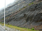 Pound Gap where the base of the visible portion this road cut marks the top the Lower Mississippian Sunbury Shale that is locally coffee colored. Most of the photograph is of the Mississippian Grainger Formation which is composed of alternating relatively continuous to occasionally shallowly channeled beds of sand with uniform sharp bedding planes and shale; both lithologies vary in thickness and the sands often exhibit Bouma sequences suggesting deposition from turbidite currents that were active over a mid to outer fan setting. The lower portion of the formation is shale rich and probably from a deeper water setting and a more distal portion of the fan. The top of the photograph in the low cloud is the overlying Lower Mississippian Newman Limestone Formation