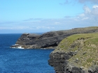 Cliffs and eroded bedding planes of the coastal monocline of Namurian deepwater sediments of the Ross Formation, overlain by turf covered Pleistocene drift. Fisherman's Point ledge to left and north. In foreground to left are bedded and slumped sediments of the Ross Formation. Turf covered Pleistocene drift exposed in foreground to right.