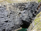 The Ross Formation of Loop Head is here expressed as turbidite sheet sands that accumulated as deepwater fan lobes that were dissected by sparse shallowly incised channels. As Elliot (2000) records these are the thicker bedded, high net-to-gross, sheet turbidites of the lower part of the Ross Sandstone Formation turbidite system. The height of these cliffs is between 45-50 m. Note the shaley partings that may compartmentalize these sands and seperate them in terms of their reservoir quality from those above. Shallow though the channeling may be it may enhance vertical reservoir continuity between the stacked sheets. Note monoclinic fold on the east side of the outcrop